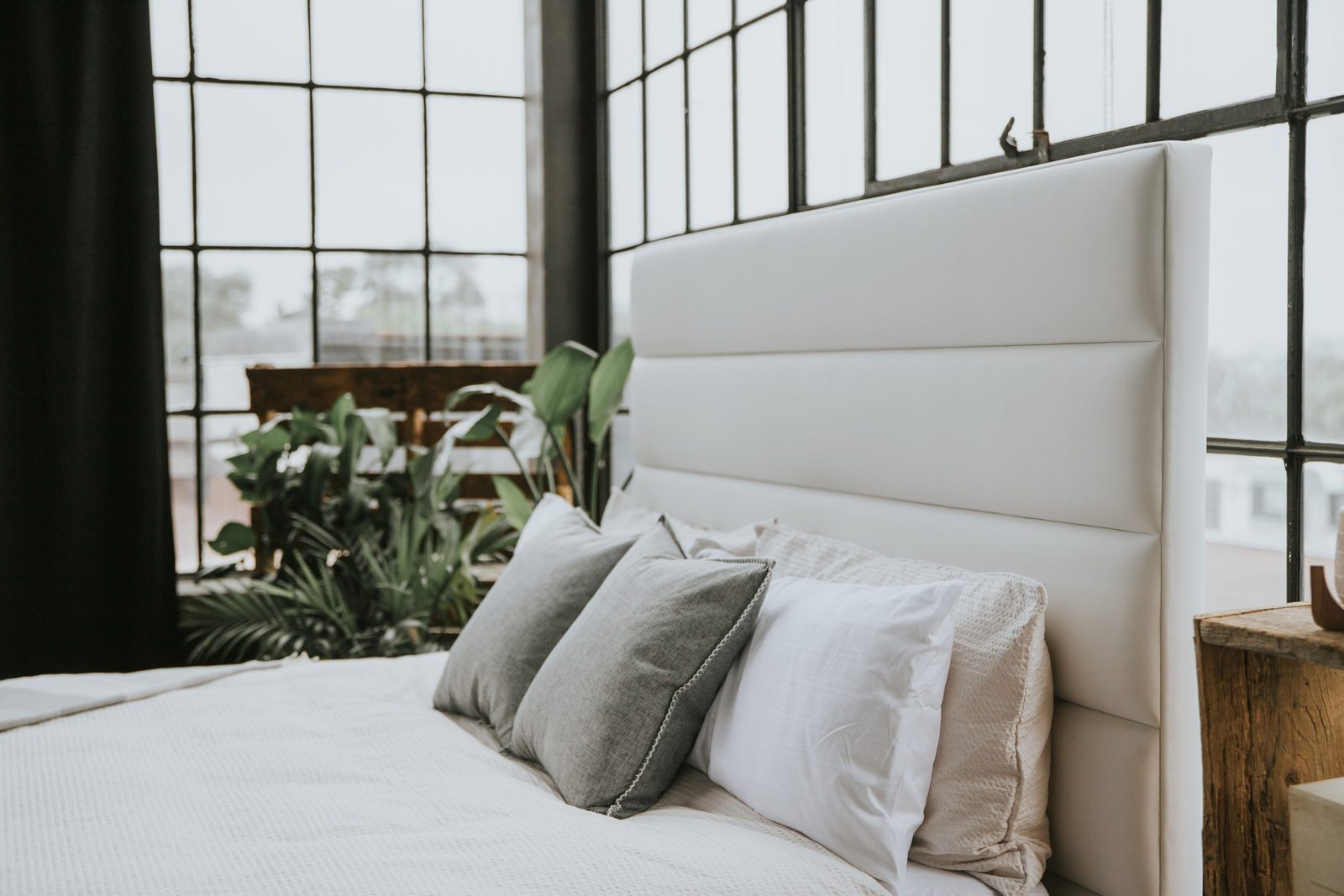 Layered grey and white pillows and a textured waffle-weave coverlet on a white upholstered Maple Zen bed in front of tall loft windows